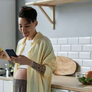 Pregnant young woman holding smartphone in kitchen smiling