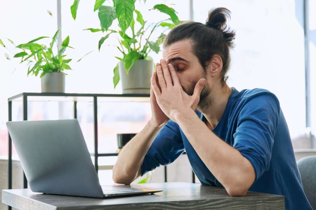 Young tired man working with laptop computer having headache fatigue